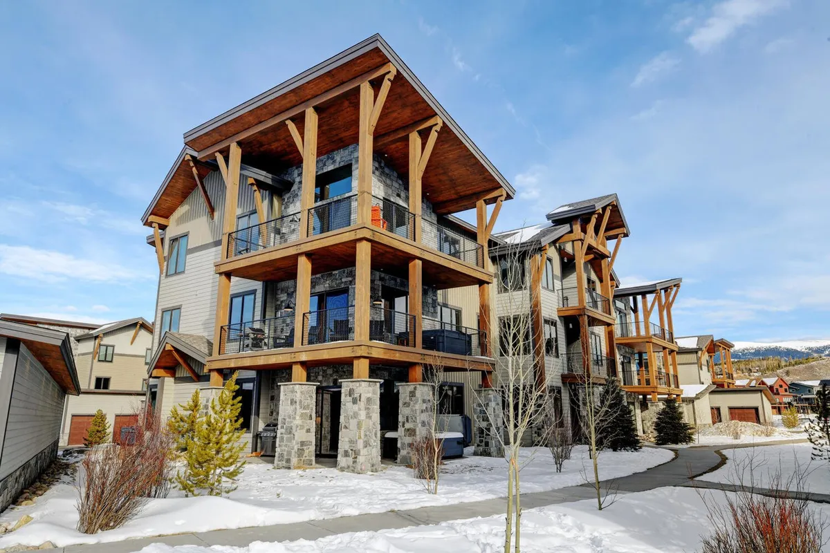 The image showcases a striking three-story luxury condo set against a snow-covered landscape in Fraser, Colorado. Large windows, wooden beams, and stone accents give the building a warm, mountain-modern charm. Balconies invite guests to enjoy the fresh alpine air, while the yard and fire pit area promise cozy gatherings and winter play.