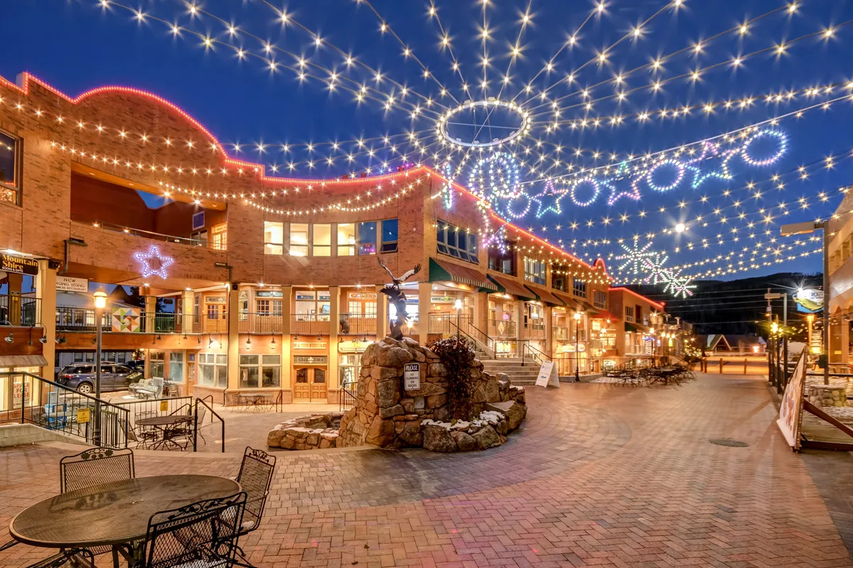 A lively exterior scene featuring decorative lights strung overhead, brick-paved walkways, charming building facades, and an inviting outdoor seating area. The space buzzes with activity, ideal for shopping, dining, and socializing during any season. The festive lighting and open layout hint at seasonal events, local markets, and evening gatherings.