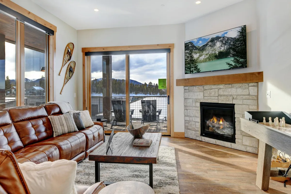 A cozy living room featuring a leather sofa, a glowing fireplace, a large wooden table, and expansive windows that offer sweeping views of the mountains. The arrangement invites relaxation and group gatherings, with the warmth of the fire and the comfort of plush seating central to the space.