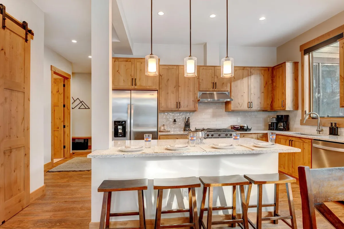 A sunlit kitchen with a central island featuring stools, stainless steel appliances, rich wooden cabinetry, and a large window that brings in natural light. The design balances functionality and warmth, emphasizing the space's appeal for both cooking and entertaining guests.