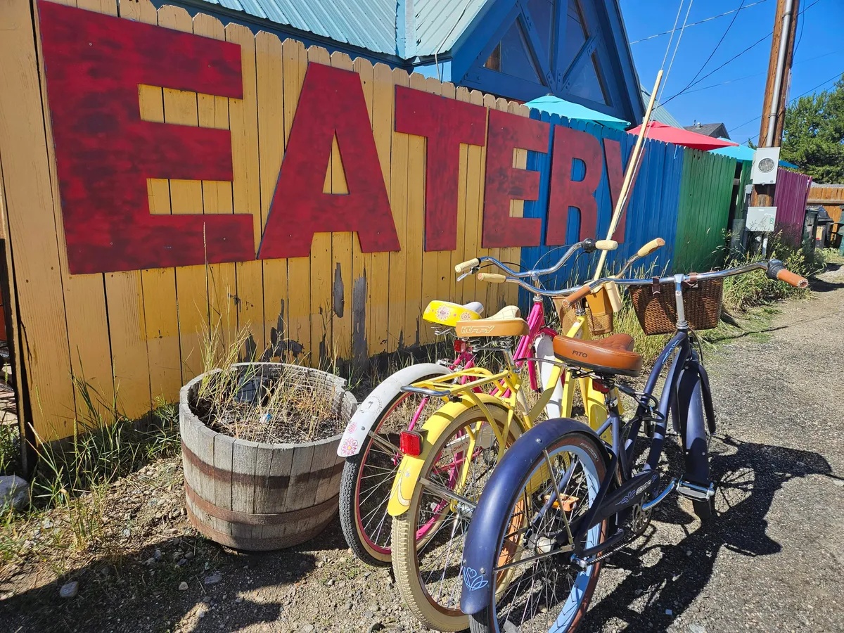 A playful exterior scene featuring a large, colorful 'EATERY' sign, vintage bicycles, and wooden barrel planters. The image suggests outdoor dining, a lively atmosphere, and the spirit of neighborhood exploration, tying into the theme of casual group outings to places like The Foundry for bowling, pizza, and movies.