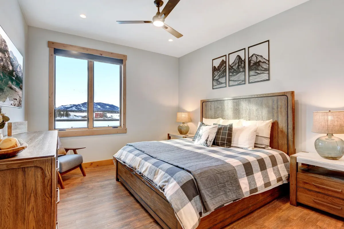 This image features a bed adorned with plaid bedding, a large window offering mountain views, wall art, and elegant light fixtures. The space invokes a sense of tranquility, comfort, and a direct connection with nature, making it an inviting place for rest and relaxation.