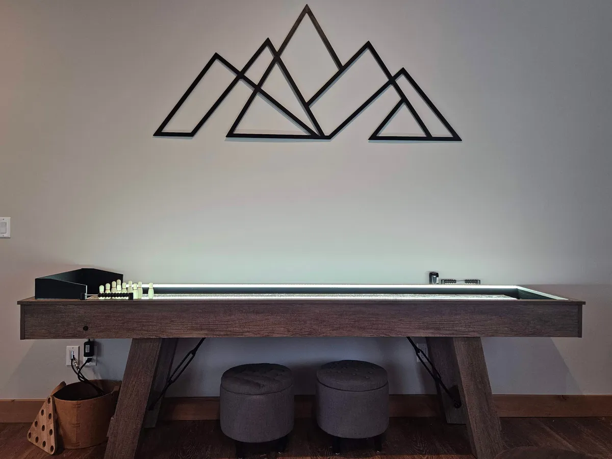 A relaxed entertainment nook equipped with a comfortable seating area and an illuminated game table, set against the backdrop of geometric mountain wall art. The scene suggests evenings spent playing board games or engaging in friendly competition.